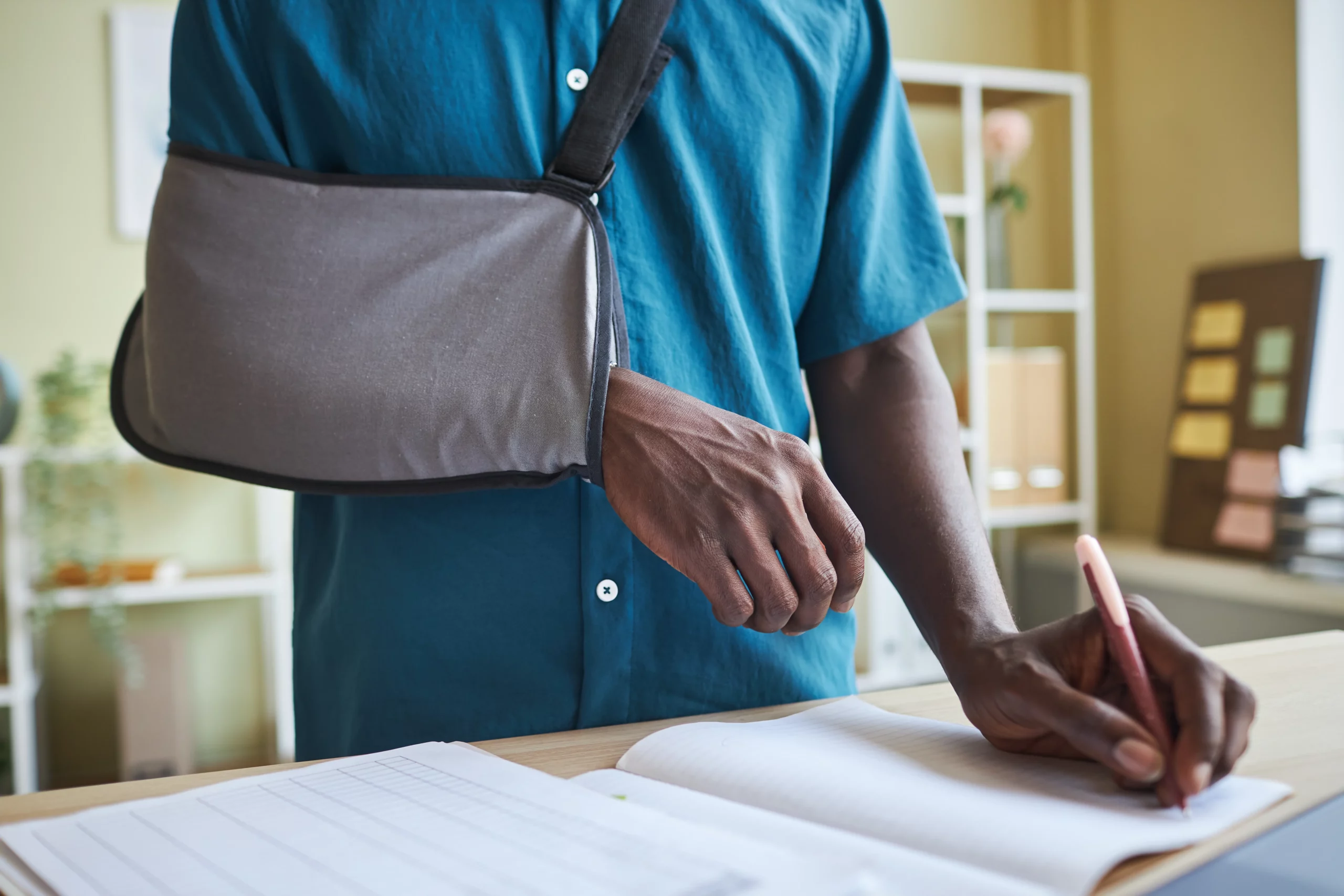 Injured person with arm sling writing notes at a desk, representing documentation and evaluation of a slip and fall injury claim and potential settlement value in Alabama